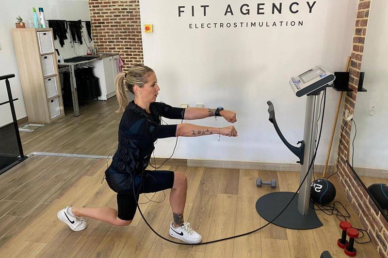 Fente en tenue d'électrostimulation devant le mur Fit Agency du studio